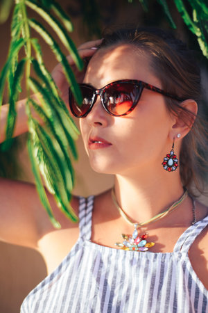 Portrait of an attractive girl in sunglasses hiding behind tree branches on a bright Sunny day,...