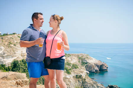 A young couple in love stands on top of a cliff overlooking a turquoise Bay with yachts, enjoying the views in their tourist adventure, sipping a cocktail from a clear glass.の写真素材