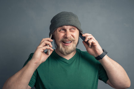 Portrait of a young man in a woolen hat talking on two phones at the same time.
