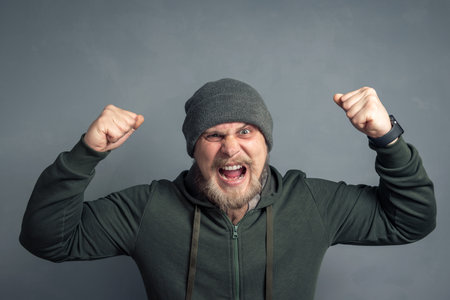 Portrait of a bearded guy in a hoodie and hat with his hands raised and clenched fists shouting Yes. The concept of luck, leadership, and winning.の写真素材
