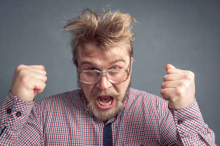 A portrait of a bearded guy with glasses and a tousled hairdo with his hands up and fists clenched and yelling Yes. The concept of luck, leadership and victory, success.の写真素材