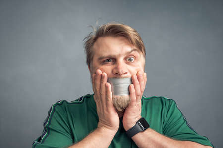 A close-up portrait of a bearded man with his mouth taped shut, holding his hands over his face in horror. The concept of what can't be said is censorship.の写真素材