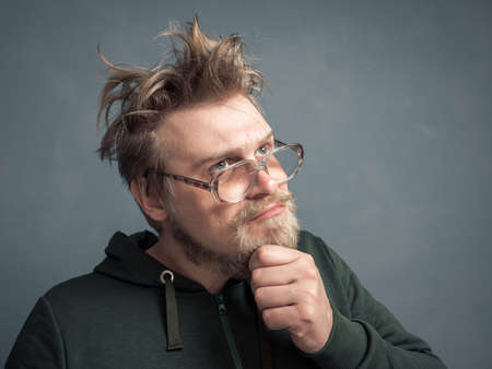 A portrait of a bearded nerd with a disheveled hairdo, gazing thoughtfully through his large glasses, scratching his chin.の写真素材