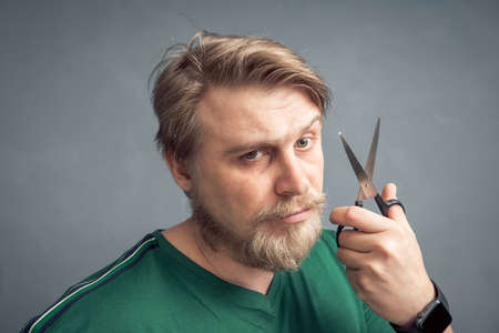 Close-up of a bearded blond man with a pair of scissors in his hand. The concept of self-care for the hair style change and hair salon.の写真素材