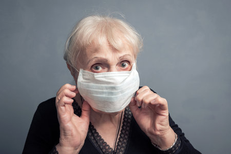 An elderly woman in a medical mask, on a gray background. Studio portrait of an adult looking at the...