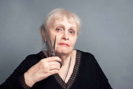 An elderly woman with a pair of scissors in her hand on a gray background. Barber services. The job of a hairdresser with sharp scissors. The concept of a Barber shop. Women's haircut.の写真素材