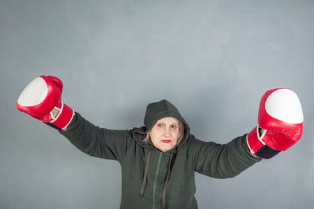 An elderly woman in Boxing gloves on a gray background. The concept of fighting, winning, fighting spirit, sports.の写真素材