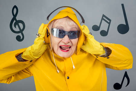 A fashionable elderly lady in yellow is listening to music with headphones. The concept of a party, a disco, a young state of mind. Close-up on a gray background.の写真素材