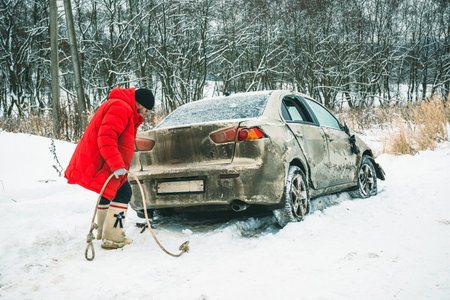 A man with a tow rope near a car wrecked on the side of the road in winter.の写真素材
