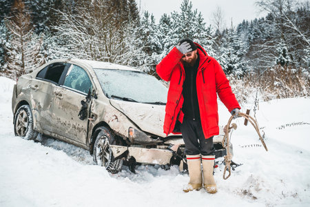 A man with a tow rope holds his head with his hand, after an accident, near a car wrecked on the side of the road in winter.の写真素材
