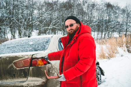 A man after an accident holds pieces of a broken car pointing at the rear marker light, on the side of the road in winter.の写真素材