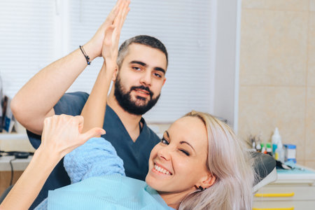A smiling blonde girl in a dental chair claps her hands with the doctor and gives a thumbs up.の写真素材