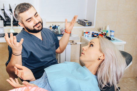 The blonde girl in the dental chair spreads her hands with the doctor discussing the treatmentの写真素材