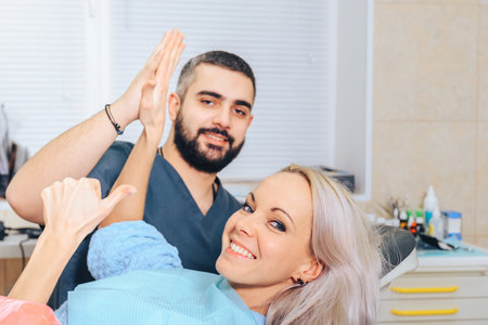 A smiling blonde girl in a dental chair claps her hands with the doctor and gives a thumbs up.の写真素材