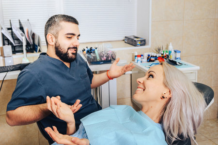 A blonde girl in a dental chair smiles and spreads her hands with a cheerful doctor discussing the treatment.の写真素材