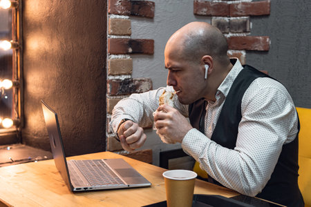 man young businessman entrepreneur caucasian,cheerful enthusiast, positive, bald in vest, sitting at a table, working laptop,looking at his watch and eats kebab, in a cafe .concept lunch time .の写真素材