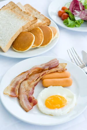 set of American breakfast including fried egg, sausage and bacon serves with salad, bread and pancakes on white tableの写真素材