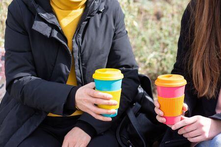 two girls sitting outdoors holding coffee cup on a city street background, take away coffee.Breakfast on the go.Sustainable lifestyle concept.reusable travel mug. Zero waste. の写真素材