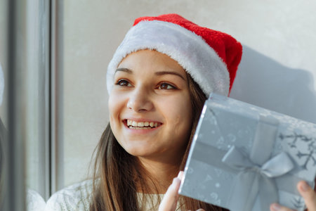 laughing girl in a Christmas hat with a gift box by the window close-up