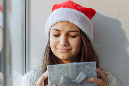 ad girl in a Christmas hat with a gift box by the window close-upの写真素材