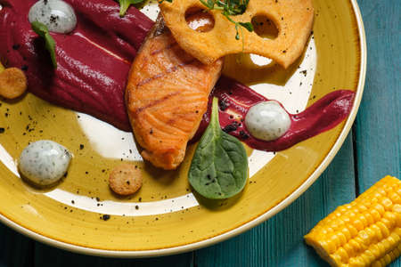 Baked salmon fillet with tomato sauce and cream sauce in bags decorated with greens and edible chips on an orange plate standing on a blue backgroundの写真素材