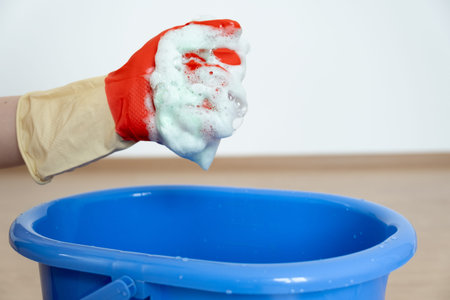A sponge with a detergent is foamed in a hand over a blue bucket.