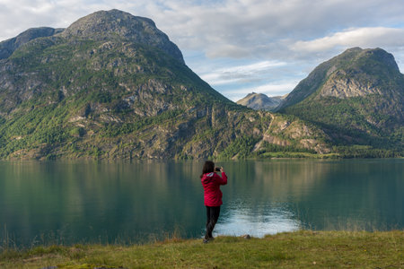 Young caucasian woman in a red jacket   taking a photo with her smartphone in a beautiful scene, on the shore of a lake, with a view towards mountainsの写真素材