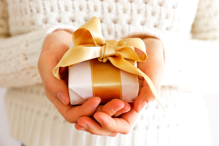 Cropped shot of woman's hands wearing white knitted wool sweater, giving present wrapped in...