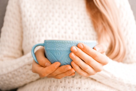 Close up young woman's hands holding big cup of hot beverage. Female wearing fashionable oversized white knitted sweater, sitting home with mug of coffee. Background, copy space.の写真素材