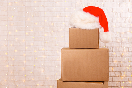 Blank brown freight box with Santa Claus hat on top, brick wall with Christmas lights on background. Moving company / delivery service holiday deals promotion concept. Copy space, close up.の写真素材