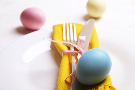 Easter table setting composition w/ pastel color eggs on plate, fork & knife silverware on white stucco plaster table background. Happy Easter greeting card. Close up, top view, copy spaceの写真素材