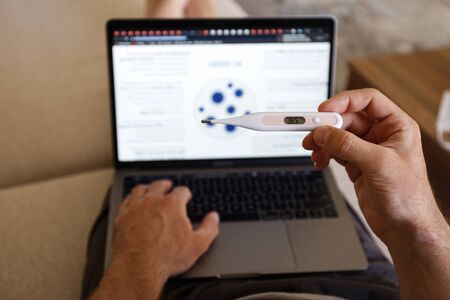 Close up shot of adult man working at home due to coronavirus quarantine concept. Male sitting on couch with laptop, measuring body temperature via digital thermometer. Background, copy space.の写真素材