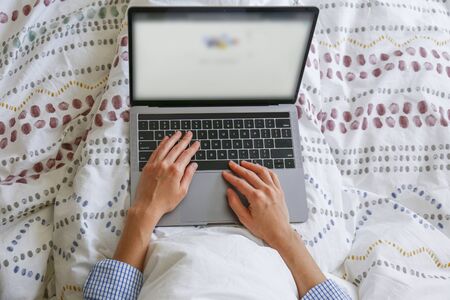 Close up shot of young blonde woman working remotely at home due to coronavirus quarantine concept. Beautiful female sitting in bed with laptop. Background, copy space.の写真素材