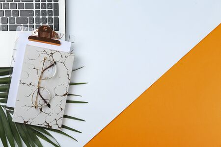 Creative designer's workplace concept. Clipboard with blank paper sheet, notebook and laptop on textured background with palm leaf. Various stationery items. Close up, copy space, top view, flat lay.の写真素材