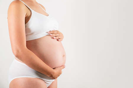 Close up of pregnant woman wearing supportive seamless maternity bra and maxi bottoms, arms on her belly. Female hands wrapped around big bare tummy. Child expectancy concept. Background, copy spaceの写真素材