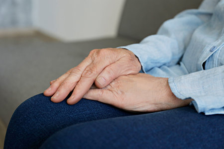 Elderly woman sitting alone at home, with hands folded on her laps. Senior old lady experiencing...
