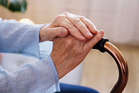 Elderly woman sitting in nursing home room holding walking quad cane with wrinkled hand. Old lady...