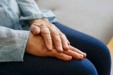 Elderly woman sitting alone at home, with hands folded on her laps. Senior old lady experiencing severe arthritis rheumatics pains, massaging, warming up arm. Close up, copy space, backgroundの写真素材