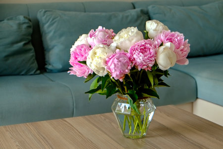 Close up shot of wooden coffee table with bouquet of beautiful white peony flowers in glass vase on foreground and blue textile couch on the background. Copy space for text, close up.の写真素材