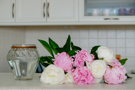 Beautiful peony flowers and a diamond shaped glass vase on a table over kitchen furniture background with a lot of copy space for text. Feminine floral composition. Close up, backdrop.の写真素材