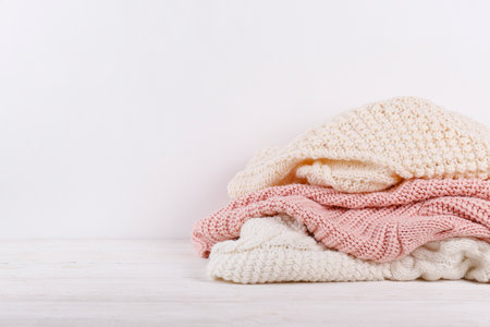 Bunch of knitted warm pastel color sweaters with different knitting patterns stacked in messy pile on white wooden table, blank wall background. Fall winter season knitwear. Close up, copy space.の写真素材