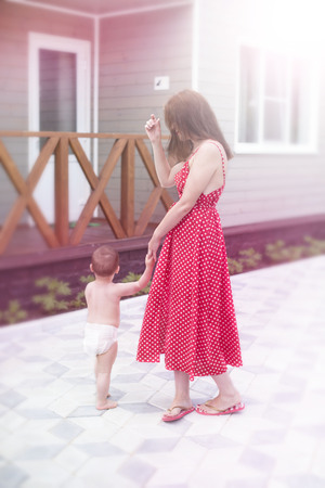 Young woman mom walking with baby son walking on the street joy happy smiling. Brunette in red dress sunlight lifestyle blurred background copy spaceの写真素材