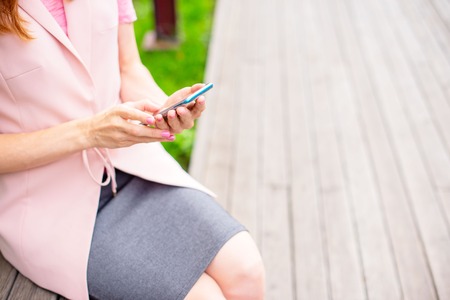 Beautiful young girl sitting on a wooden bench in the open Looking at the mobile phone Womens hands Sunny day green bushes lifestyle Selective focus copy spaceの写真素材