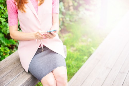 Beautiful young girl sitting on a wooden bench in the open Looking at the mobile phone. Women's hands Sunny day green bushes lifestyle Selective focus copy spaceの写真素材