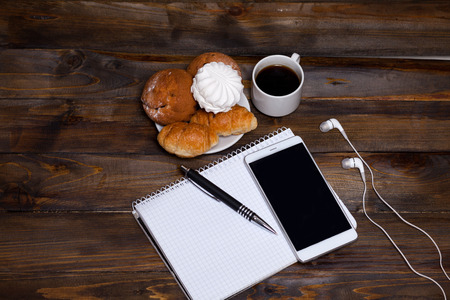 White cup of coffee with headphones and mobile phone, and lying next to notebook with pen, croissant, marshmallow on wooden backgroundの写真素材