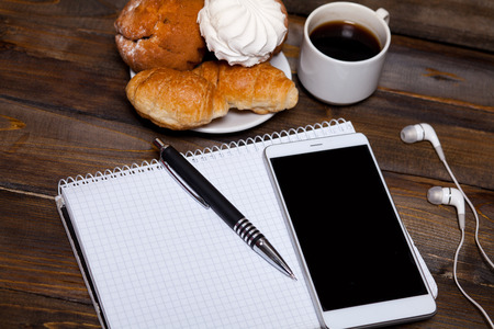 White cup of coffee with headphones and mobile phone, and lying next to notebook with pen, croissant, marshmallow on wooden backgroundの写真素材