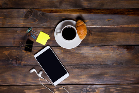 White mobile phone on wooden wooden background with headphones, cup of coffee, croissant and stationery appliances top viewの写真素材