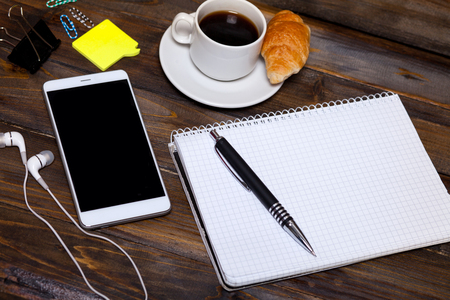 White mobile phone on wooden wooden background with headphones, cup of coffee, croissant and Stationery appliances.の写真素材