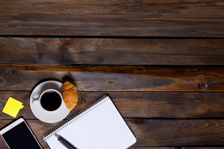 White mobile phone on wooden wooden background with headphones, cup of coffee, croissant and Stationery appliances top viewの写真素材