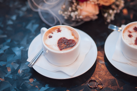 Two white cappuccino cups with hearts and wedding rings, bride bouquet on the table.の写真素材